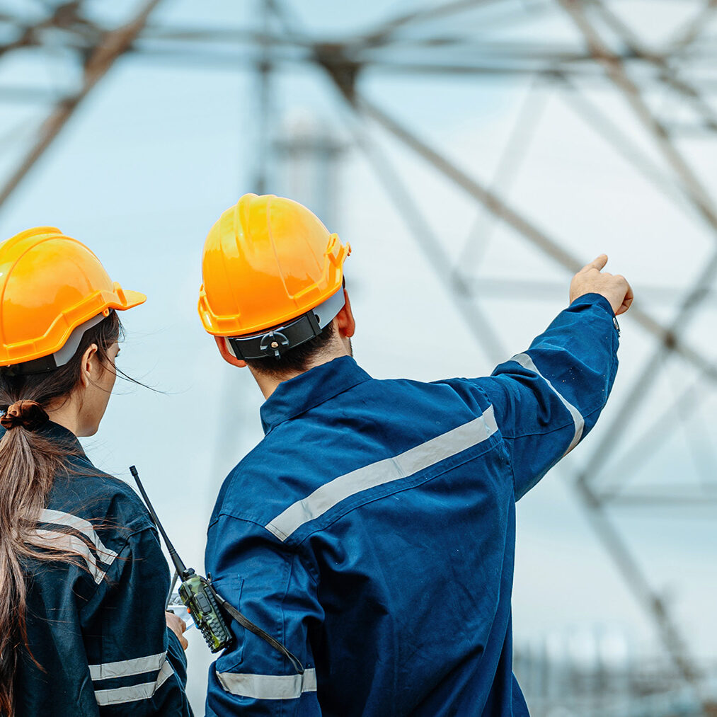 Engineers inspect power lines and discuss safety protocols at electrical substation site during daytime Two engineers wearing safety helmets and uniforms are discussing maintenance tasks at an electrical substation. They are focused on the structure and ensuring safety measures.